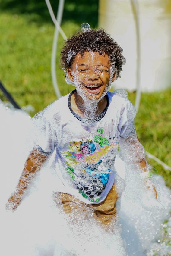 A child laughing, covered in foam, at an outdoor community event.