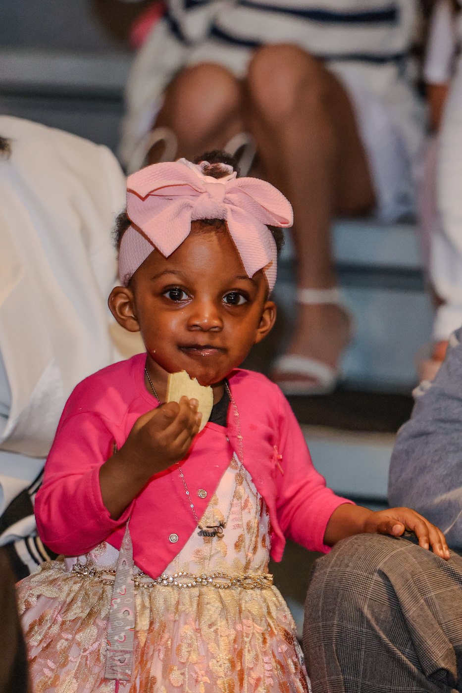 A toddler in a pink bow watches from the audience, snack in hand.